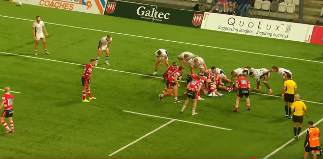 Rugby union match in progress at Kingsholm Stadium showing players from two teams - one in red and white hooped jerseys, the other in white - engaged in a scrum. QuoLux advertising board reading 'Supporting leaders. Inspiring success.' is prominently displayed along the pitch perimeter.